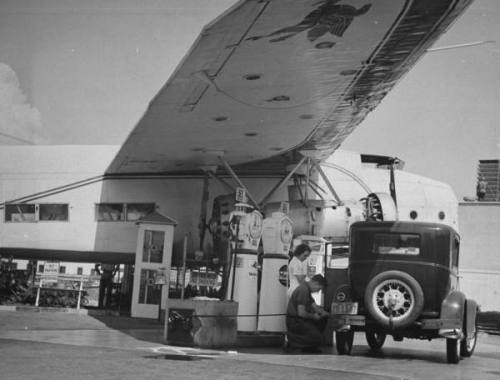 Service attendant pumping gasoline into Ford sedan as woman watches at gas pumps covered by the wing of a large permanently parked airplane, on Wilshire Boulevard --circa 1938.