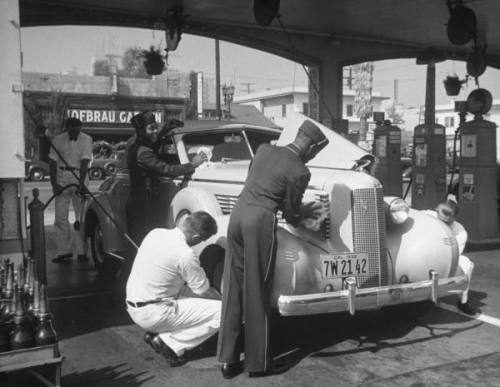 Muller Brothers Service Station's white uniformed attendents pumping gas & inflating tires on a fancy convertible while their uniformed African-Amer. conterparts wield rags as they polish the windshield & chrome --ca. 1938.